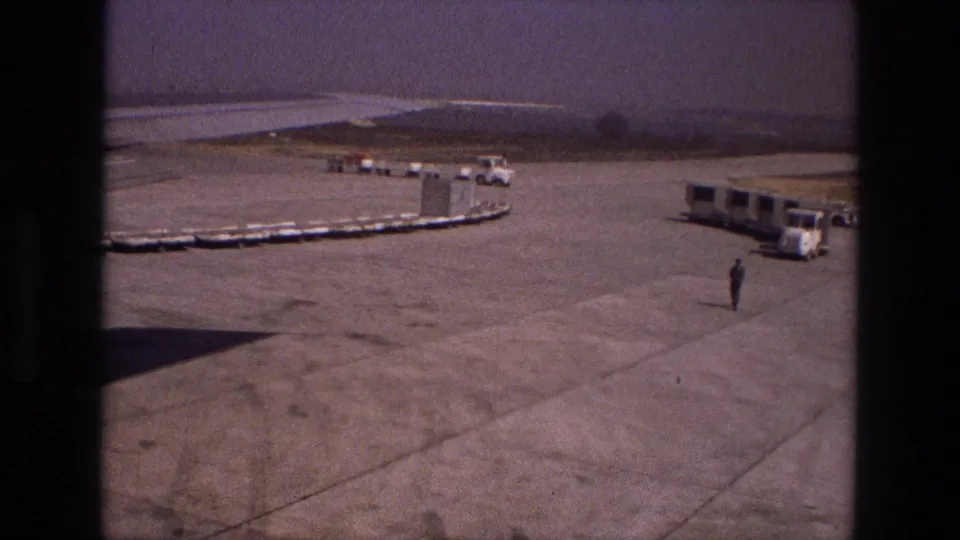 A handheld 8mm view of an Iranian airport tarmac in 1972, showing luggage carts, a passenger bus, distant vehicles, and a solitary worker.