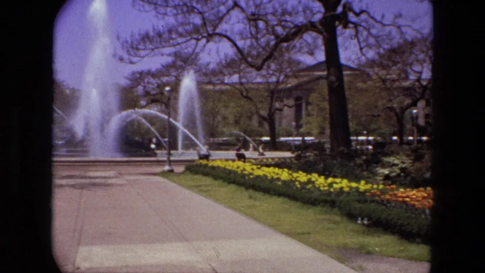 1972 Iran park frame showing a multi-jet fountain, concrete path, tulip beds, trees, passing cars, and a small distant person near the road.