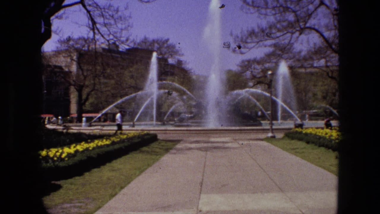 1972 Iran 8mm frame with high fountain spray, arcing side jets, trimmed lawn, yellow flowers, trees, and small distant people.