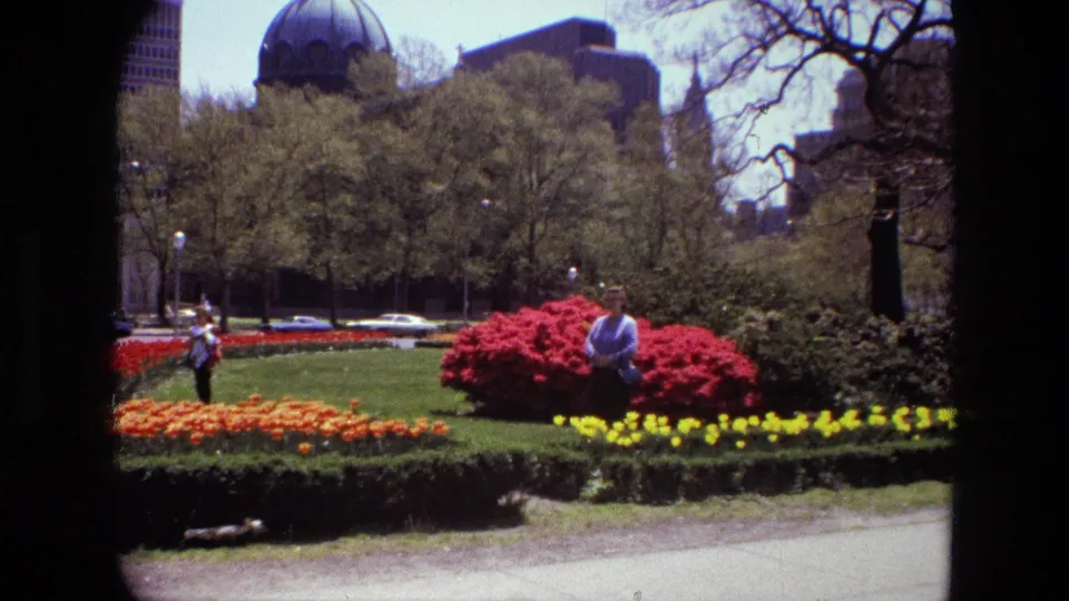 1972 Iran 8mm frame showing bright flower beds, a green lawn, pedestrians, parked cars, trees, and urban buildings behind the park.