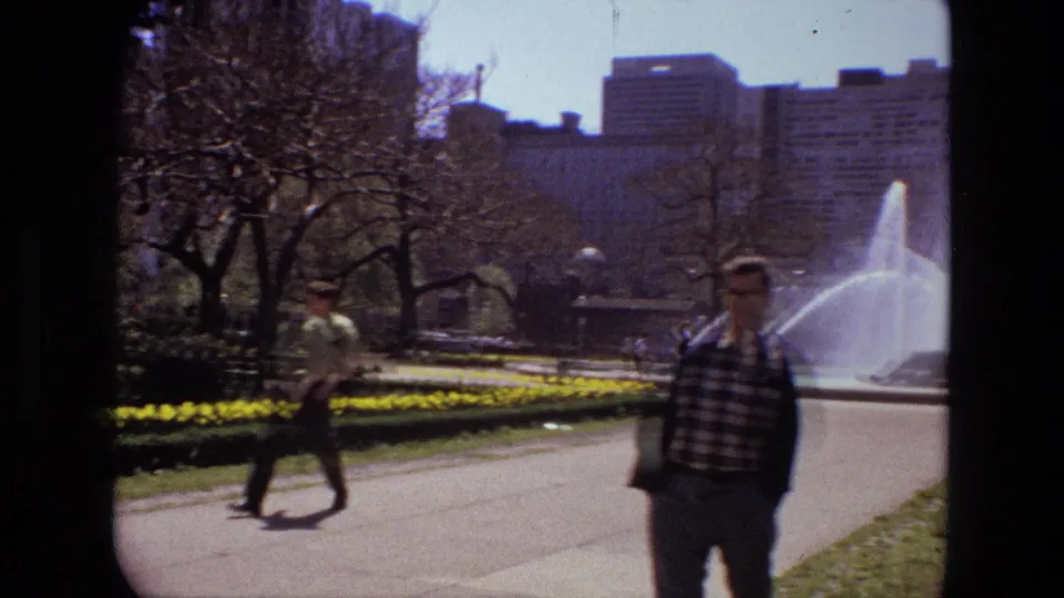 1972 Iran park frame with pedestrians in the foreground, a fountain spraying water, yellow flowers, bare trees, and urban buildings behind.