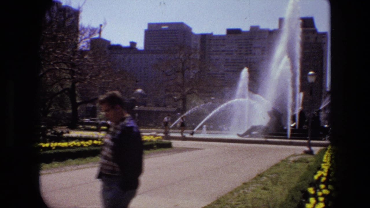 Pedestrian passes a public fountain and flower beds in a sunny garden.