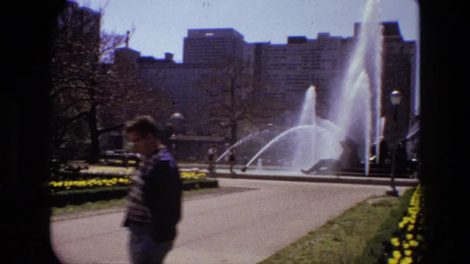 In 1972 Iran, a man walks past an active fountain, paths, lawns, and flower beds in a public garden.