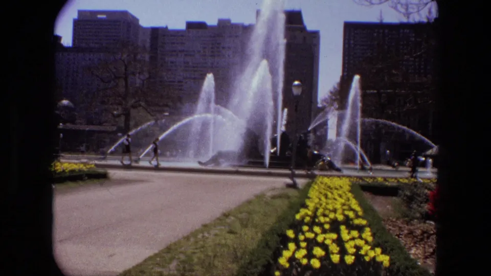 A landscaped public garden in 1972 Iran shows arcing fountain jets, yellow flower beds, lawns, and distant buildings.