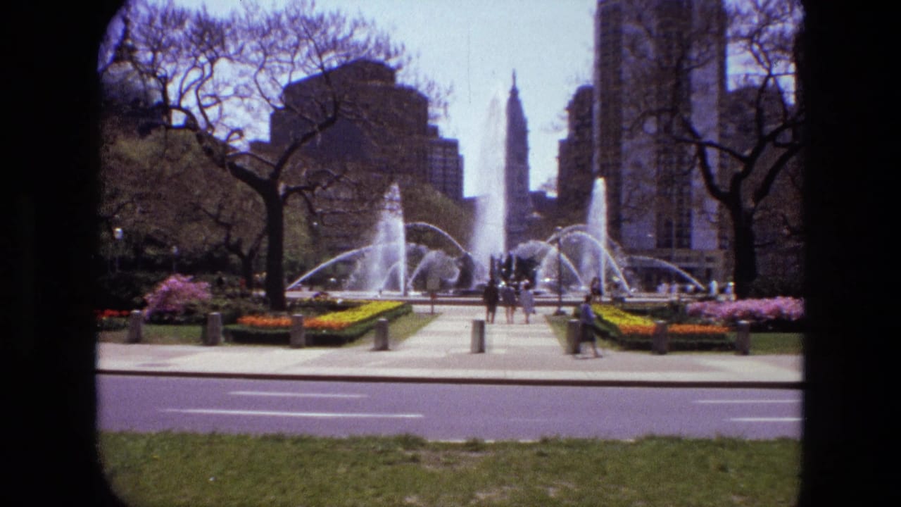 Large fountain plaza with flowerbeds, pedestrians, trees, road, and tall buildings.