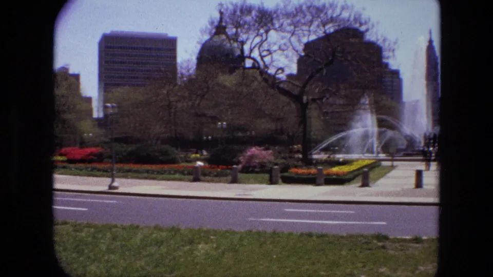 1972 Iran home-movie view across a street toward a fountain, spring flowerbeds, trees, and modern buildings around a public square.