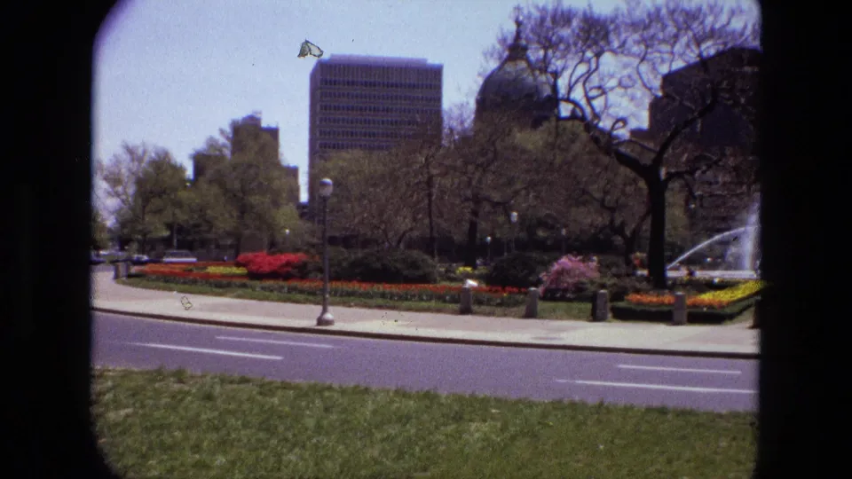 Wide 1972 Iran frame showing a landscaped square with grass, colorful planting, trees, road, buildings, and part of the fountain at right.