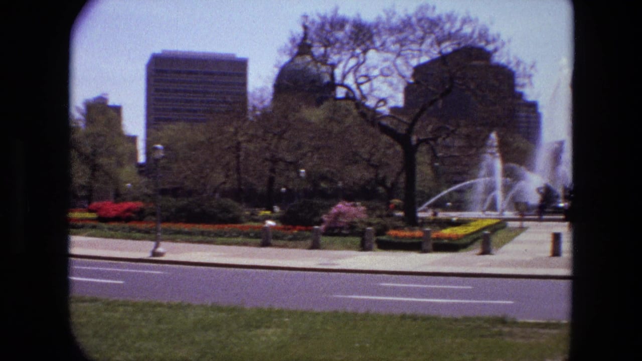 Urban park fountain across a road with flower beds, trees, and high-rise buildings behind it.