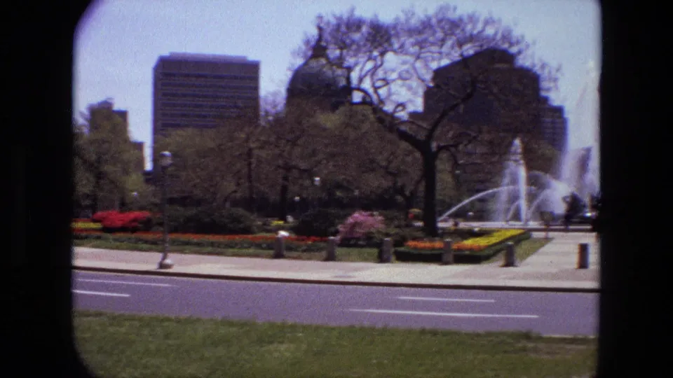 1972 Iran home-movie frame showing a city park fountain, landscaped beds, bare trees, and high-rise buildings across a road.