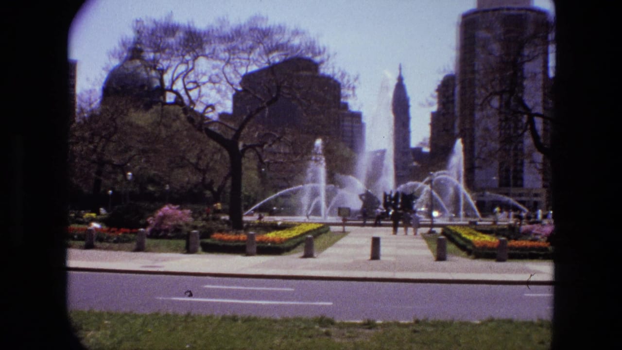 Park fountain with pedestrians, flower beds, trees, and tall city buildings in the background.