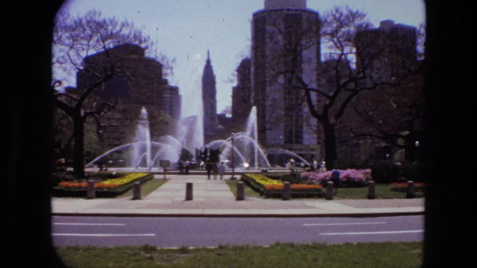 1972 Iran home-movie view of a fountain plaza with pedestrians, colorful planting beds, trees, and urban towers.
