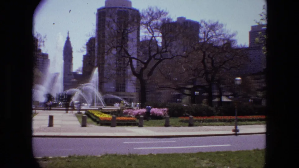 1972 Iran frame showing a landscaped metropolitan park fountain, distant people, roadway, trees, and high-rise buildings.