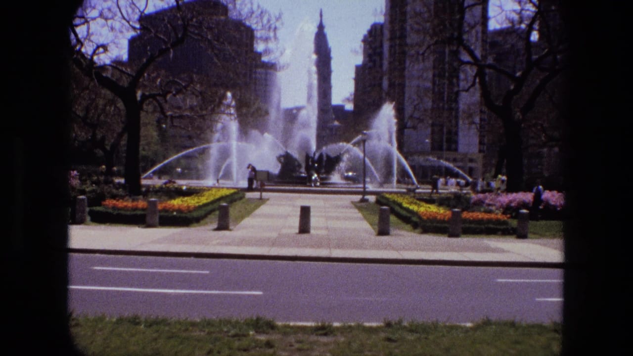 1972 Iran urban park scene with arcing fountain jets, paved walks, flowerbeds, pedestrians, trees, and high-rise buildings.