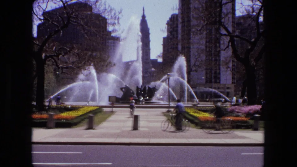 1972 Iran frame showing a cyclist on the sidewalk in front of a large fountain, with pedestrians, flowers, trees, and buildings behind.
