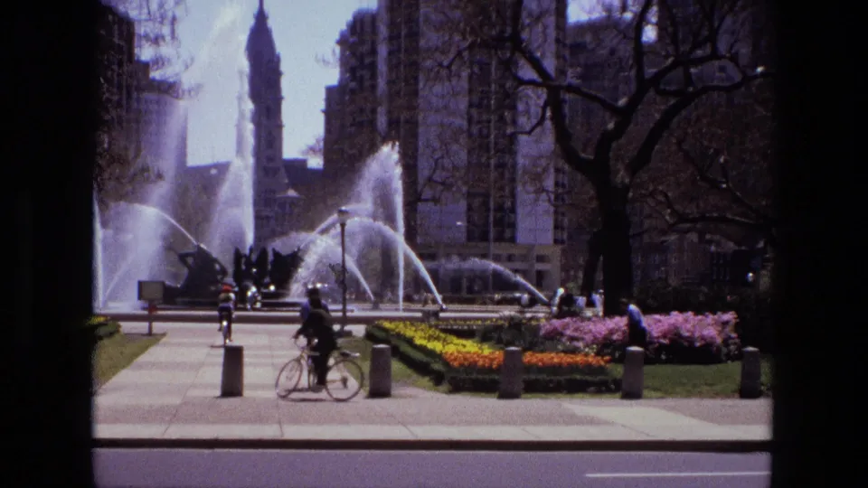 1972 Iran home-movie still with a bicyclist crossing the foreground, fountain jets behind, flowerbeds, pedestrians, trees, and city buildings.