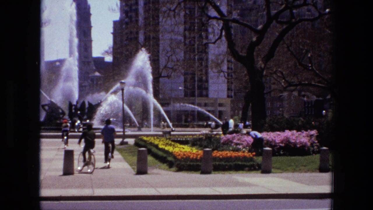 Urban park with fountain spray, flower beds, cyclists, pedestrians, trees, and tall buildings.