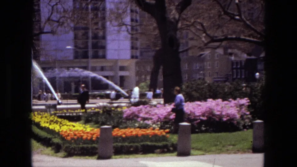 1972 Iran 8mm frame of landscaped flower beds, fountain spray, a pedestrian, trees, bollards, and urban buildings beyond the park.