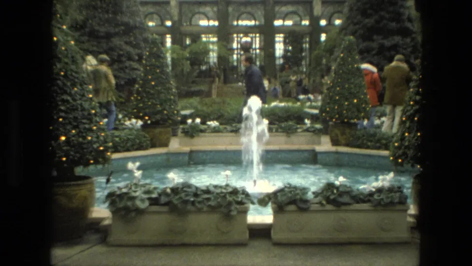 1972 Iran 8mm frame showing a fountain pool, decorated evergreen trees, stone planters, and visitors in winter coats inside an indoor garden.