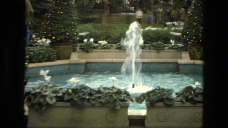 1972 Iran 8mm frame centered on a tall fountain jet, blue pool water, white flowers, and decorated greenery in an indoor garden.
