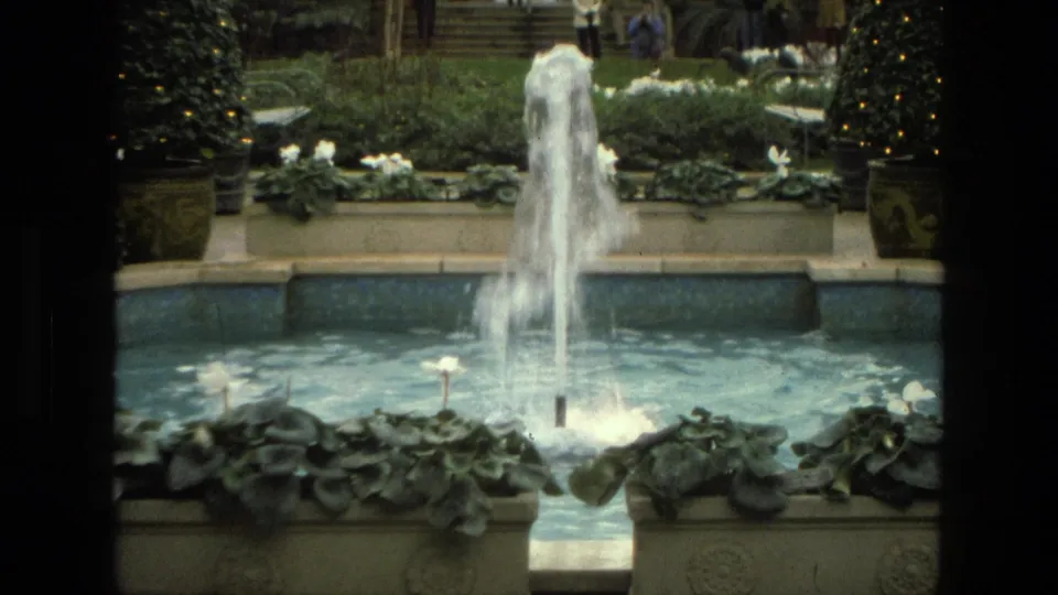 1972 Iran 8mm frame showing the fountain closer in view, with water spray, blue pool water, leafy planters, and a decorated tree at the edge.