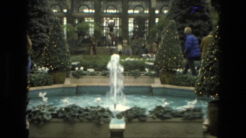1972 Iran 8mm frame showing a fountain, stone planters, decorated evergreen trees, arched glass windows, and visitors moving through the garden.
