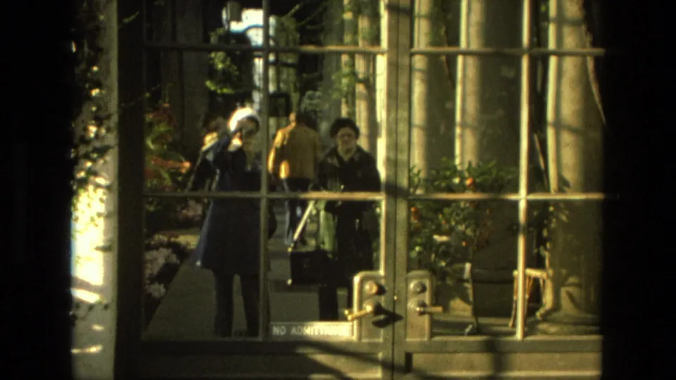 1972 Iran 8mm frame looking through greenhouse glass at several visitors walking near plants and metal door handles.