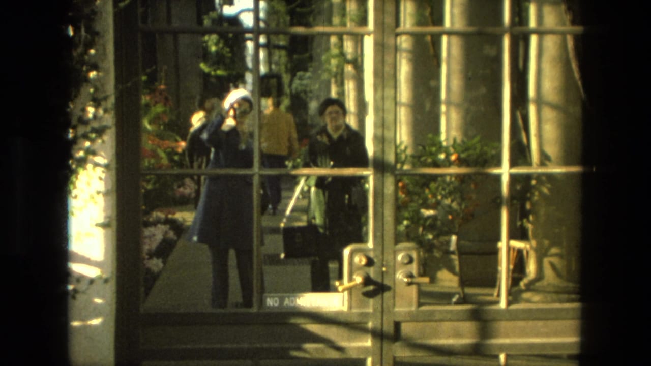 Two adults stand beyond glass doors near pink flowers and greenery.