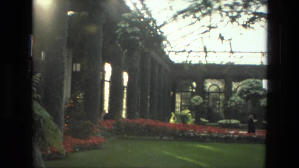 1972 Iran 8mm frame looking down a greenhouse path toward red flower beds, glass roofing, arched windows, and a distant walking visitor.