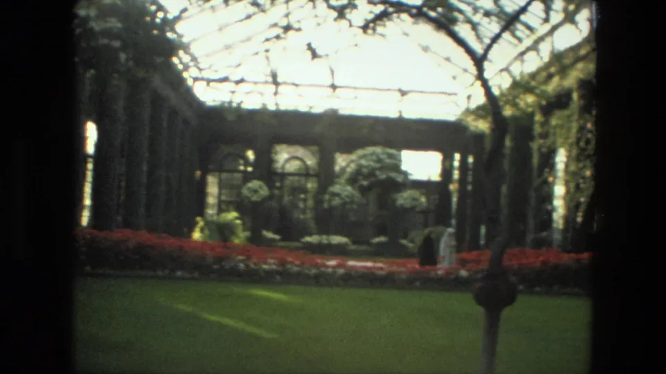 1972 Iran home-movie frame showing a greenhouse garden with red flower beds, glass roof, arched windows, and a visitor walking near the camera.