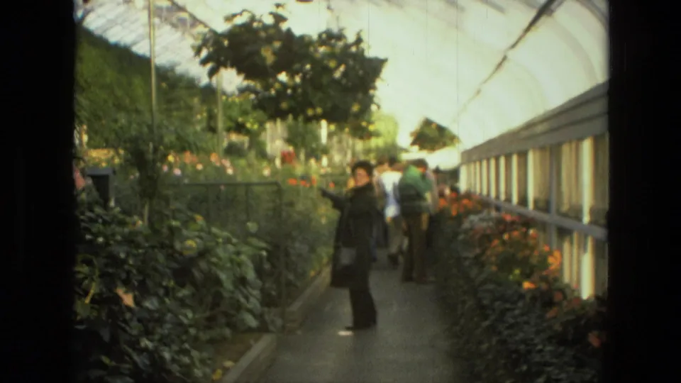 1972 Iran frame showing an adult woman near the camera in a greenhouse corridor with flowers, foliage, visitors, and glass panels.