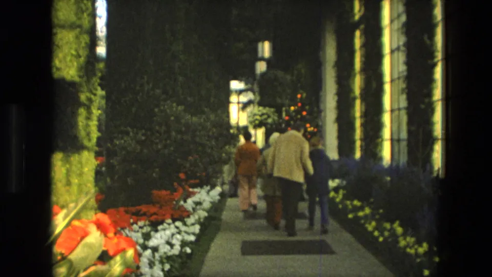Visitors in a 1972 Iran 8mm home movie walk through a warm-lit indoor garden with flower beds and tall greenery.