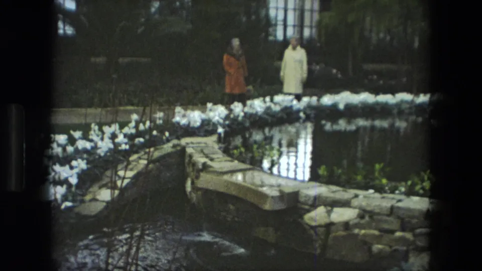 Two women in period coats walk beside a pond in Iran, with white flowers, stone borders, and dark garden greenery visible.