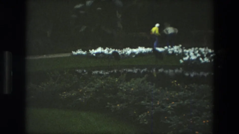 1972 Iran 8mm frame showing a manicured garden lawn, white flower beds, shrubs, and a distant person walking through the grounds.