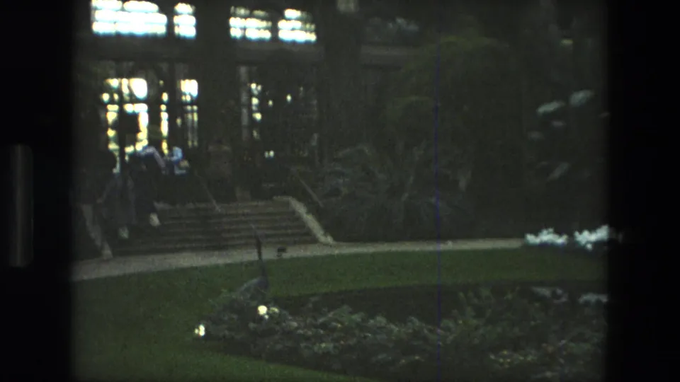 1972 Iran 8mm frame showing garden steps, lawns, planted beds, and distant people near a large building with lit windows.