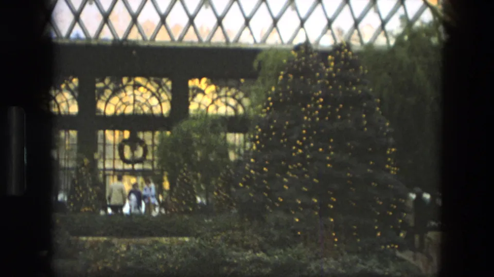 Soft 8mm view of a decorated indoor garden in Iran, with a large wreath, warm tree lights, shrubs, and visitors near the glass wall.