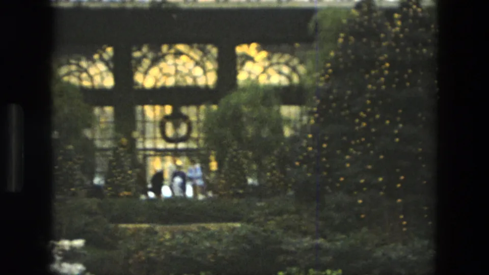 1972 Iran frame showing visitors in a conservatory garden, with illuminated holiday trees, a hanging wreath, and soft home-movie exposure.