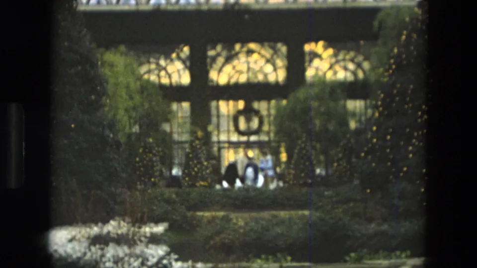 1972 Iran home-movie frame showing planted indoor garden beds below patterned glass windows, with distant visitors partly visible.