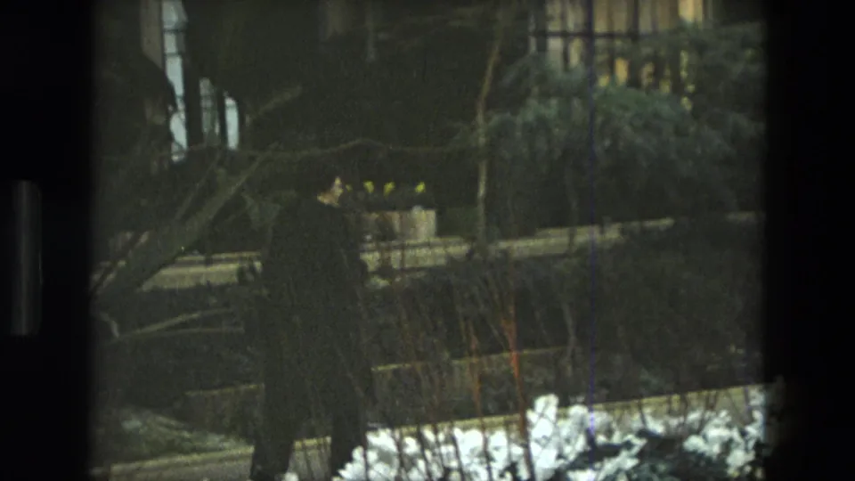 1972 Iran 8mm frame with a dark figure walking past white flower beds, foliage and low garden structures at night.