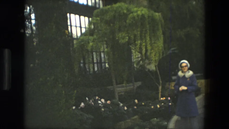 Adult woman in a blue coat and white headscarf stands near the edge of the frame in a 1972 Iranian garden scene.