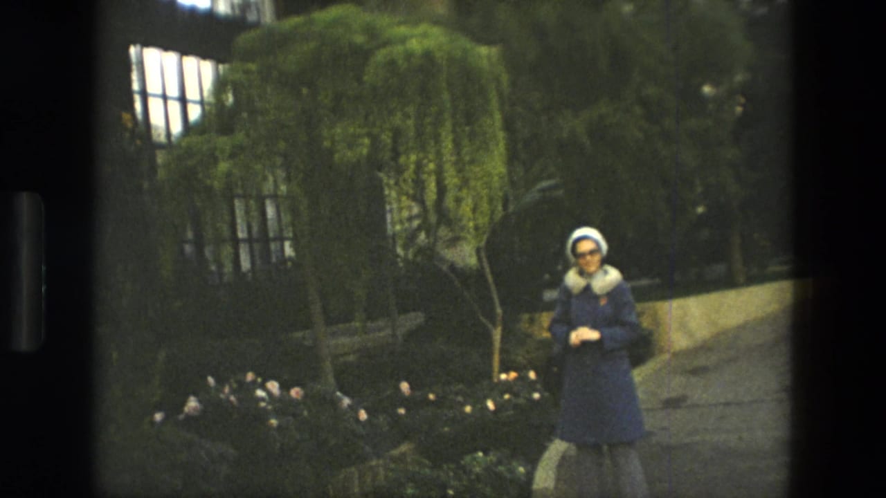 Woman in a blue coat and white hat standing on a garden path near plants.