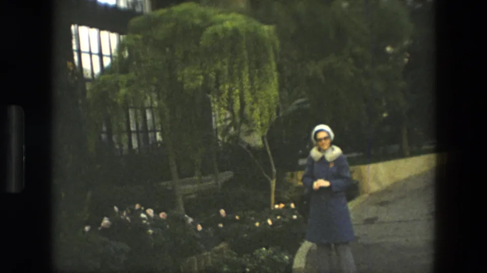 In 1972 Iran home-movie footage, a woman in a blue coat and white hat stands on a path among greenery and flower beds.