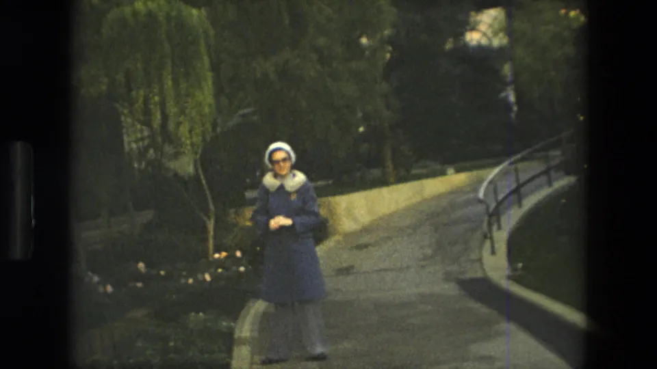 A woman in a blue coat and white hat faces the camera on a curved garden walkway in Iran, 1972, with greenery behind her.