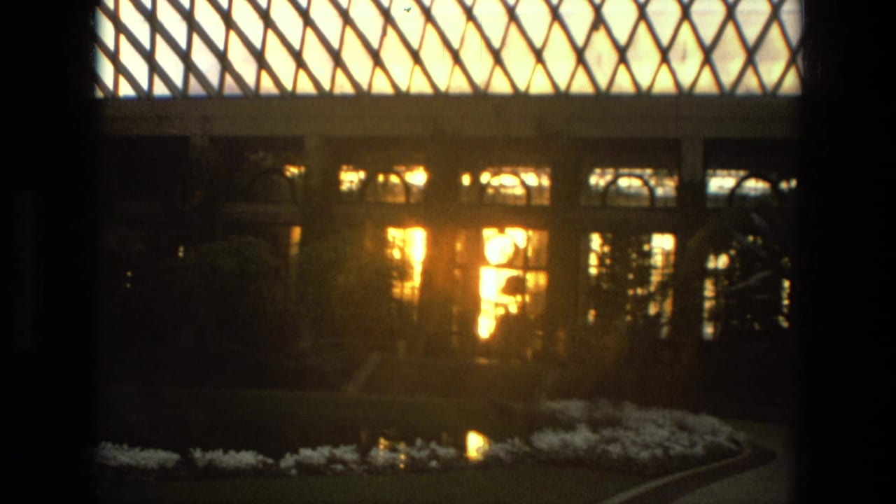 1972 Iran 8mm frame of a glass-roofed garden facade with arched windows, bright interior light, shrubs, flower beds, and reflective water.