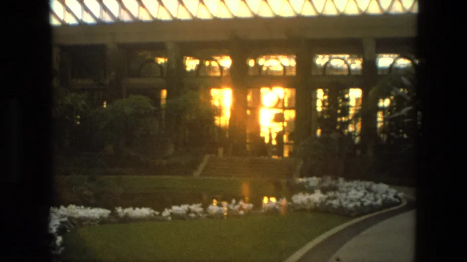1972 Iran 8mm frame showing a manicured garden path, lawn, white flower bed, pond reflections, and a glowing glass-and-window facade.