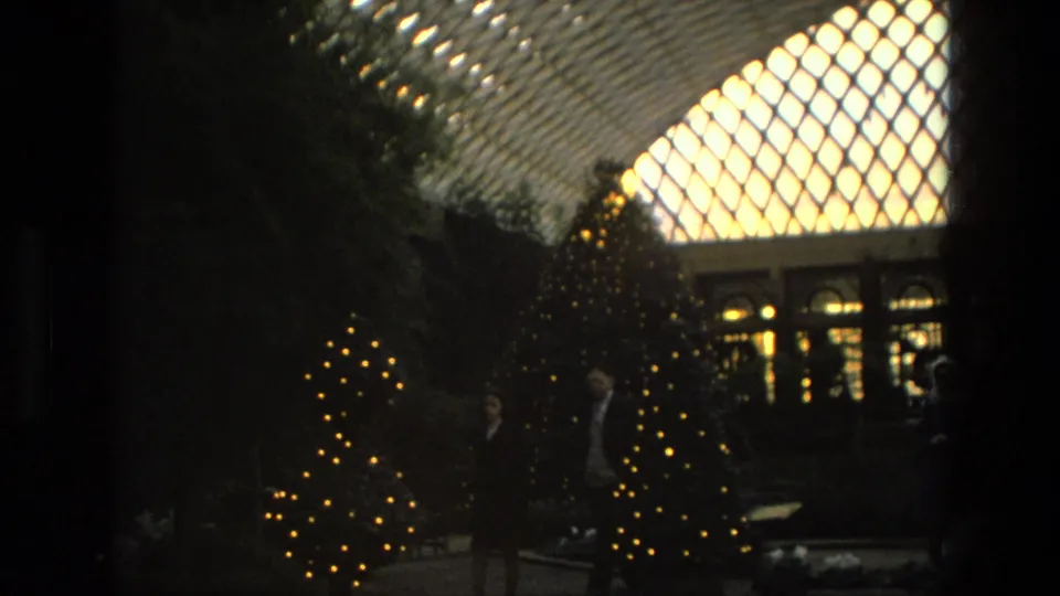 1972 Iran home-movie frame showing lit Christmas trees, shadowed visitors, plants and a glowing arched glass roof.