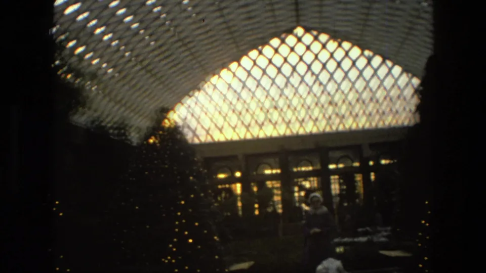 1972 Iran home-movie still showing a hatted visitor, fountain or basin, lit trees and the bright patterned glass ceiling.