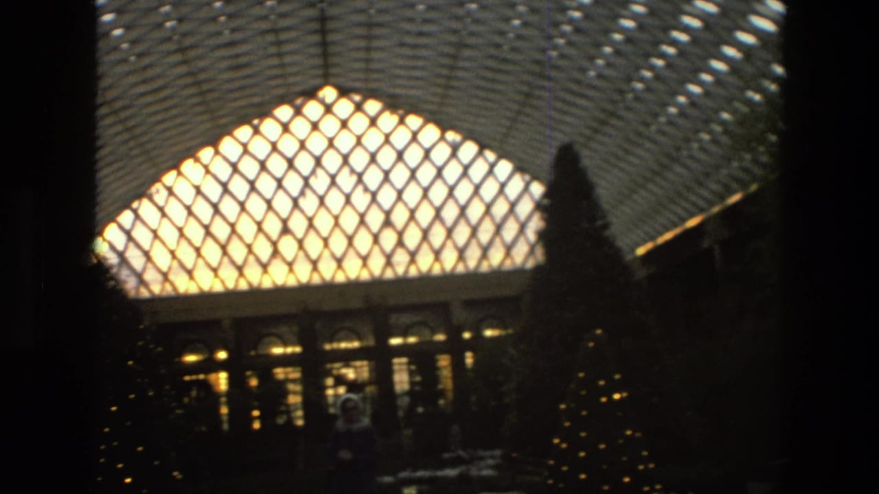 1972 Iran home-movie frame with a bright lattice roof, dark indoor trees, warm windows, and faint figures in silhouette.