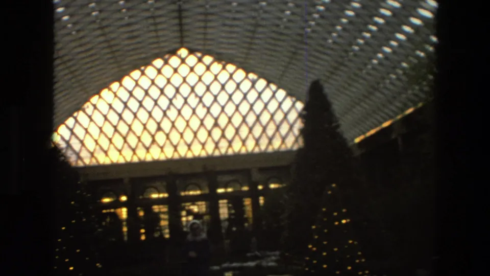 1972 Iran home-movie frame with a bright lattice roof, dark indoor trees, warm windows, and faint figures in silhouette.