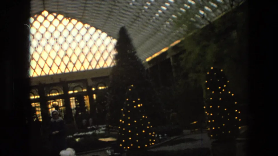 1972 Iran footage frame with illuminated Christmas trees, arched windows, shrubs, and a distant visitor under a bright glass roof.