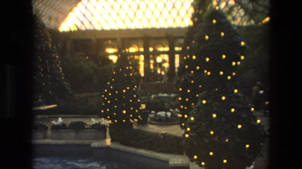 1972 Iran home-movie view of lit Christmas trees around a fountain, with clipped plantings and bright patterned glazing overhead.
